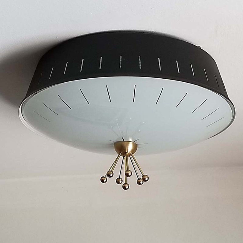 Może przedstawiać: Lampa sufitowa z matowym czarnym metalowym kloszem w kształcie bębna z pionowymi wycięciami. Sp&oacute;d jest zakryty matowym białym szklanym dyfuzorem. Centralny mosiężny element zwieńczający rozciąga się w d&oacute;ł z sześcioma promienistymi szprychami.