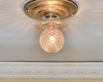 May include: A vintage ceiling light fixture with a gold-toned metal base and a decorative, clear glass globe. The globe has a diamond pattern and is illuminated, casting a warm glow. The fixture is mounted on a white ceiling.
