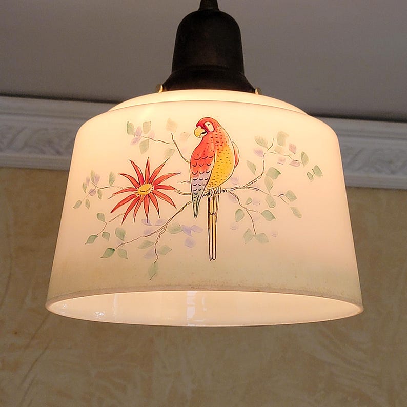 May include: A white glass lampshade with a painted design of a red, yellow, and green parrot perched on a branch with a red flower. The lampshade is hanging from a black metal fixture.