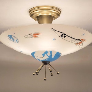 May include: A vintage-style ceiling light fixture with a white, saucer-shaped shade. The shade features illustrations of rockets, planets, and a blue and white globe. The fixture has a brass-colored base and decorative arms with spherical accents.