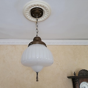 May include: A white glass pendant light fixture with a brass chain and a decorative brass ceiling plate. The glass shade has a ribbed design and a small tassel hanging from the bottom.