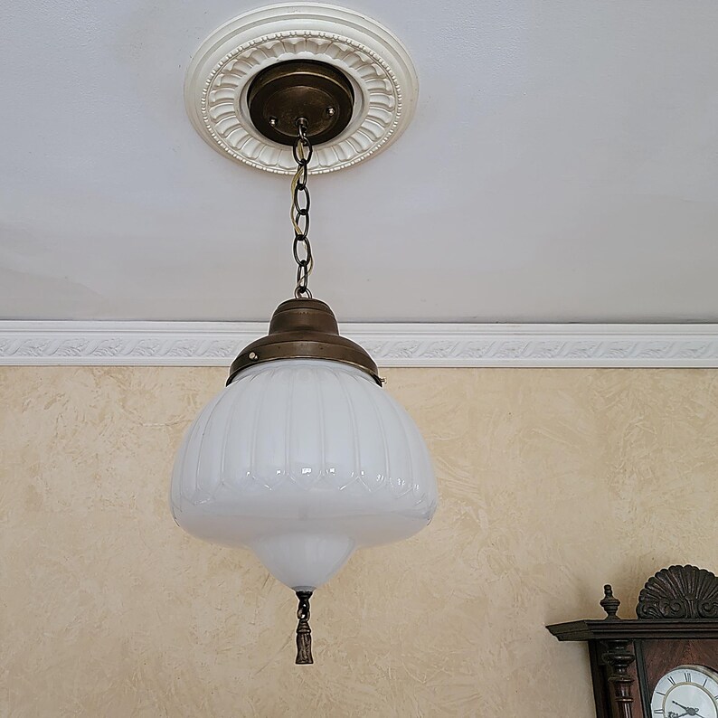 May include: A white glass pendant light fixture with a brass chain and a decorative brass ceiling plate. The glass shade has a ribbed design and a small tassel hanging from the bottom.