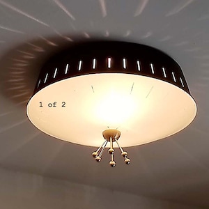 May include: A flush mount ceiling light fixture with a dark brown drum shade featuring vertical cutouts. The interior is white, and the center features a gold-toned metal finial with four radiating arms ending in small spheres. Text reads 1 of 2.