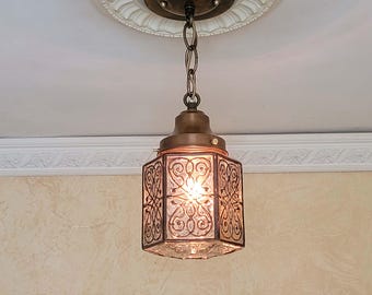 Może przedstawiać: Wisząca lampa wisząca z sześciokątnym szklanym kloszem ozdobionym misternym, ciemnym metalowym filigranem. Oprawa jest zawieszona na pasującym ciemnym metalowym łańcuchu przymocowanym do okrągłej podsufitki z dekoracyjną białą sztukaterią.