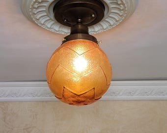 May include: A ceiling-mounted light fitting featuring a spherical, textured amber glass shade with a repeating chevron pattern. The shade is secured by a dark bronze-toned metal base attached to a white, circular, decorative ceiling medallion.
