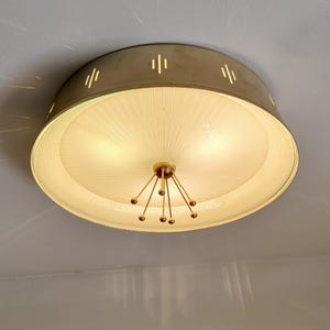 May include: A round flush mount ceiling light fixture with a metallic gold outer rim featuring vertical cutouts. The center consists of a white ribbed glass shade with a decorative gold-toned central hub featuring multiple radiating thin rods.