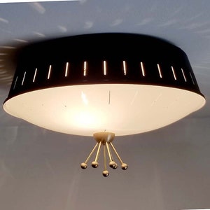Może przedstawiać: Lampa sufitowa z szerokim, ciemnobrązowym metalowym kloszem w kształcie bębna z pionowymi wycięciami. Sp&oacute;d stanowi duży, p&oacute;łprzezroczysty biały szklany dyfuzor. Centralne złote zwieńczenie z pięcioma promienistymi ramionami z kulkami.