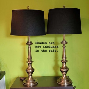 May include: A pair of gold-toned table lamps with black lampshades. The lamps have a classic design with a fluted base and a round finial. "Shades are not included in the sale." is written in black text on a white background.