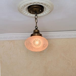 May include: A ceiling pendant light fixture featuring a brass-toned metal canopy and chain suspension. The light includes a frosted glass shade with an etched floral pattern, showcasing a central daisy-like flower surrounded by leaf motifs.