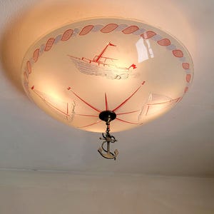 May include: A nautical-themed ceiling light fixture. The frosted glass shade features a red and white boat design, a compass, and a decorative rope border. A bronze anchor pendant hangs below the fixture.