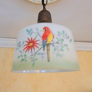 May include: A white glass lampshade with a painted design of a parrot on a branch with green leaves and a red flower. The parrot is red, yellow, and blue.