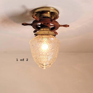 May include: A ceiling light fixture featuring a wooden ship wheel design with brass accents and a textured clear glass shade. The fixture is mounted flush to the ceiling and includes the text 1 of 2 visible on the left side of the image.