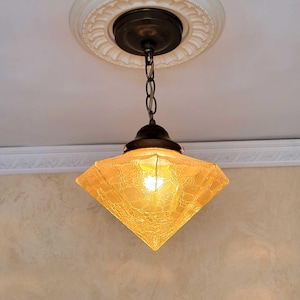 May include: A vintage-style pendant light fixture with a diamond-shaped, textured amber glass shade. The light hangs from a chain and a dark metal canopy attached to the ceiling. The ceiling has a decorative white molding.