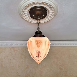 May include: A vintage-style pendant light fixture with a frosted glass shade. The shade features a peach and brown color scheme with decorative tree and scroll designs. The fixture is suspended from a bronze-colored ceiling mount.