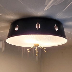 May include: A black mid-century modern flush mount ceiling light fixture. The light has a circular shade with three sets of vertical cutouts. The fixture has a gold-colored, starburst-style base with small spherical accents.