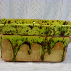 May include: A two-piece ceramic baking dish. The top is a shallow tray, the bottom a deeper dish with handles. The dishes are yellow-green with brown speckles and a dripped glaze effect. Measurements are in centimetres.