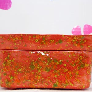 May include: A rectangular ceramic baking dish with a glossy finish, featuring a vibrant red base color speckled with orange, yellow, and green paint splatters. The dish has two side handles and a matching rectangular lid resting on top.