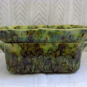 May include: A two-piece ceramic serving dish set. The set features a rectangular base with handles and a matching lid. The dishes have a mottled green and brown glaze, giving them a unique, textured appearance. The lid has a scalloped edge.