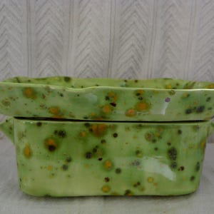 May include: A two-piece ceramic serving dish set. The set features a light green glaze with speckled orange and brown accents. The top piece is a shallow, rectangular dish with a slightly wavy edge, and the bottom piece is a rectangular container with small handles.