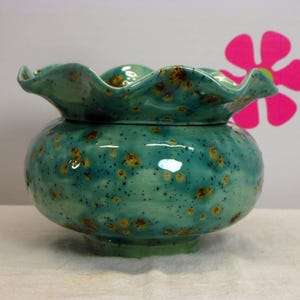 May include: A round ceramic bowl with a ruffled, wavy rim. The bowl features a glossy turquoise glaze with a speckled pattern of dark brown and golden-yellow spots distributed across the entire surface, including the flared, scalloped edge.