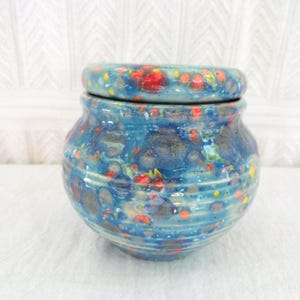 May include: A blue ceramic jar with a matching lid. The jar and lid feature a speckled pattern of red, yellow, and orange dots. The jar is approximately 13 cm tall.