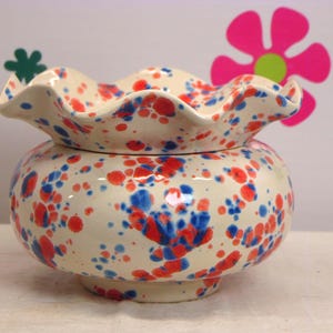 May include: A ceramic bowl with a ruffled, wavy rim and a rounded body. The surface is off-white and features a speckled pattern of vibrant red and blue paint splatters distributed across the entire exterior and the interior of the rim.