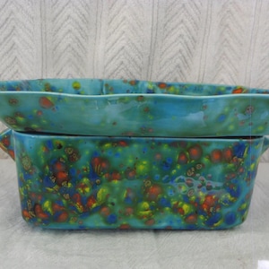 May include: Two rectangular ceramic baking dishes stacked vertically, featuring a vibrant glaze with a mottled pattern of turquoise, blue, green, yellow, and orange spots. Each dish has small integrated side handles and a smooth, glossy finish.