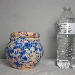 May include: A ceramic jar with a lid, featuring a speckled design in blue, orange, and yellow against a peach-colored base. The jar is next to a clear plastic water bottle.
