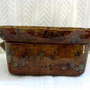 May include: A rectangular ceramic baking dish with a glossy, mottled brown glaze featuring darker brown and greenish spots. The dish has a wide, flared rim and small handles on the sides, showcasing a deep, rustic, and earthy color palette.