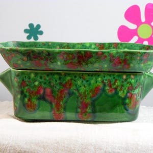 May include: A rectangular ceramic baking dish with two side handles, featuring a glossy green glaze with mottled red and dark blue drip patterns throughout the exterior and interior surfaces, showcasing a vibrant and multi-colored finish.