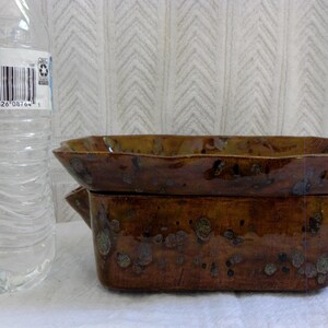 Pu&ograve; includere: Una pirofila rettangolare in ceramica marrone con coperchio abbinato. Il piatto ha una finitura lucida con accenti maculati. Il coperchio ha un bordo smerlato. A sinistra c'&egrave; una bottiglia d'acqua di plastica trasparente.