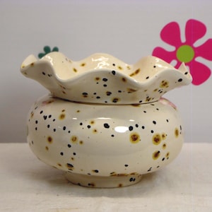 May include: A cream-coloured ceramic bowl with a wide, ruffled rim. The exterior and the interior of the rim feature a speckled pattern consisting of small black dots and larger, blurred brown spots distributed across the smooth, glossy surface.