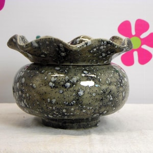 May include: A ceramic bowl with a ruffled, scalloped rim and a rounded body. The surface features a glossy, sage green glaze with a speckled pattern of dark navy blue and white dots throughout the exterior and the interior of the rim.