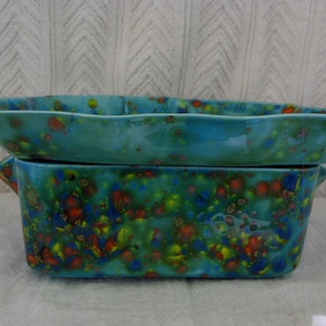 May include: Two ceramic serving dishes. The top dish is a shallow, turquoise-colored oval shape. The bottom dish is rectangular with handles and a speckled pattern of green, blue, yellow, and orange. Both dishes have a glossy finish.