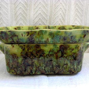 May include: A rectangular ceramic baking dish with a glossy glaze featuring a mottled pattern of lime green, dark brown, and teal. The dish has a wide, flared rim, integrated side handles, and a deep basin with a smooth, undulating top edge.