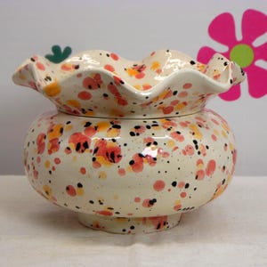 May include: A cream-coloured ceramic bowl with a ruffled, scalloped rim. The exterior and interior feature a speckled pattern of orange, pink, and black paint splatters. The bowl has a rounded, bulbous body that tapers down to a circular base.