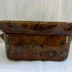 May include: A rectangular, brown ceramic baking dish with a matching lid. The dish and lid have a glossy finish with speckled, dark brown and green accents. The lid has a slightly wavy edge and sits atop the dish.