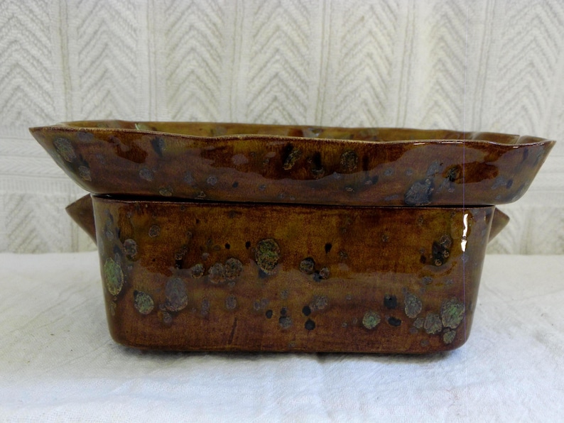 Pu&ograve; includere: Una pirofila rettangolare in ceramica marrone con coperchio abbinato. La pirofila e il coperchio hanno una finitura lucida con accenti maculati marrone scuro e verdi. Il coperchio ha un bordo leggermente ondulato.