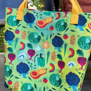 May include: A green tote bag with a colorful vegetable pattern, including artichokes, tomatoes, and carrots. The bag has yellow handles and is made of fabric. The bag is sitting on a wooden surface.