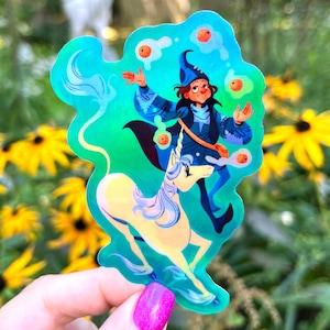 May include: A die-cut sticker with a holographic finish featuring a wizard in a blue robe juggling orange spheres above a white unicorn. The wizard has dark hair and a blue pointed hat, while the unicorn has a long flowing mane and tail.