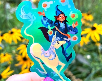 May include: A die-cut sticker with a holographic finish featuring a wizard in a blue robe juggling orange spheres above a white unicorn. The wizard has dark hair and a blue pointed hat, while the unicorn has a long flowing mane and tail.