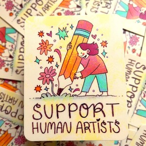 May include: A yellow and orange pencil with a blue stripe is being held by a person with pink hair. The person is wearing a pink and blue shirt and blue pants. The background is yellow with pink flowers, stars, and hearts. The text "SUPPORT HUMAN ARTISTS" is written in black at the bottom of the image.