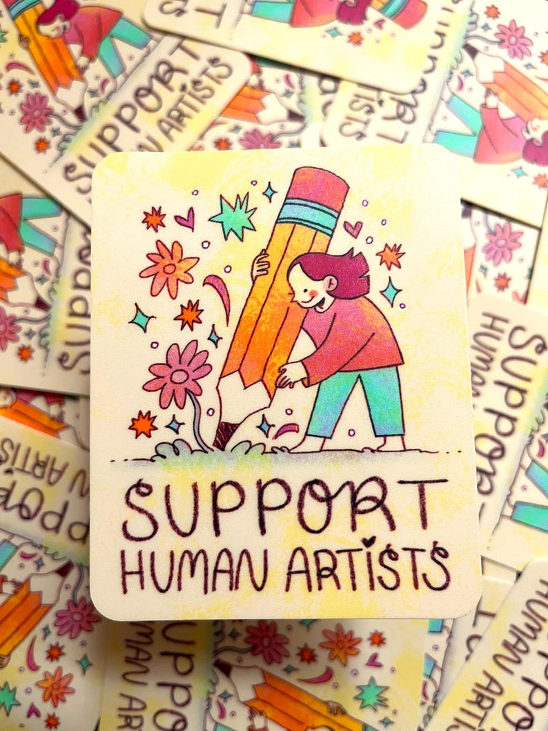 May include: A yellow and orange pencil with a blue stripe is being held by a person with pink hair. The person is wearing a pink and blue shirt and blue pants. The background is yellow with pink flowers, stars, and hearts. The text "SUPPORT HUMAN ARTISTS" is written in black at the bottom of the image.
