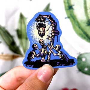 Pu&ograve; includere: Un adesivo con uno sfondo blu con tre figure scheletriche che raggiungono una lanterna luminosa con campane appese.