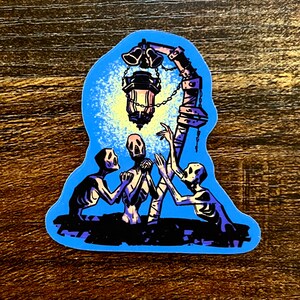 Pu&ograve; includere: Un adesivo con uno sfondo blu che mostra tre figure scheletriche che raggiungono una lanterna con campane appese. La lanterna &egrave; accesa e proietta un bagliore sulle figure.