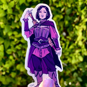 Puede incluir: Una pegatina morada y rosa de un personaje femenino con el pelo corto y oscuro, que lleva un atuendo morado y rosa y sostiene una espada. La pegatina tiene el texto "TAC" en la esquina inferior izquierda.