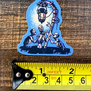 Pu&ograve; includere: Un adesivo con un'illustrazione blu e viola di tre scheletri che raggiungono una lanterna appesa a un palo con delle campane. La lanterna &egrave; accesa e proietta un bagliore sugli scheletri.
