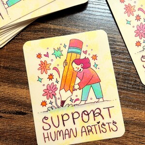 May include: A yellow card with a cartoon illustration of a person holding a large pencil. The pencil is surrounded by colorful flowers and stars. The text "SUPPORT HUMAN ARTISTS" is written in black at the bottom of the card.