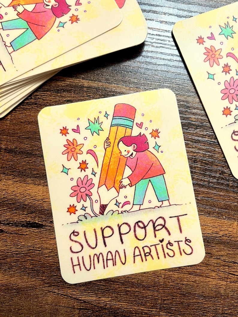 May include: A yellow card with a cartoon illustration of a person holding a large pencil. The pencil is surrounded by colorful flowers and stars. The text "SUPPORT HUMAN ARTISTS" is written in black at the bottom of the card.