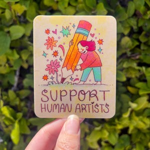 May include: A sticker with a cartoon illustration of a person holding a large pencil with the text "SUPPORT HUMAN ARTISTS" below.